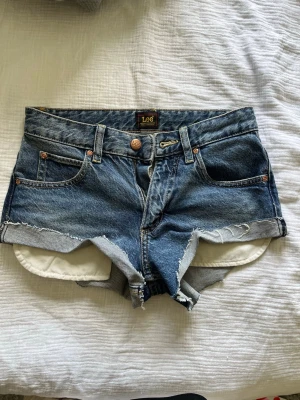 Blå jeansshorts från Lee - Snygga blå jeansshorts från Lee med råa kanter och klassisk femficksdesign. Shortsen har låg midja.