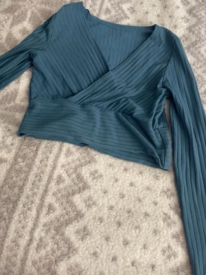 Blå ribbad omlottopp - Snygg blå croppad omlottopp med ribbad struktur och långa ärmar. Toppen har en djup v-ringning och bred midjeresår för en bekväm passform. Perfekt att styla med höga jeans eller kjol.