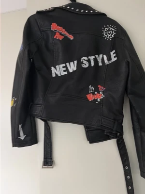 Svart skinnjacka med nitar och tryck - Cool svart skinnjacka från Pull&Bear med silvriga nitar på kragen, bälte i midjan och massor av vita och röda tryck som 'NEW STYLE', stjärnor och text. Jackan har dragkedjor, fickor och en edgy streetstil. Storlek L men mycket mindre egentligen.
