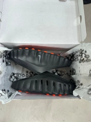 SMÅ I STORLEK  svarta tofflor med orange sula - Säljer ett par svarta-tofflor med en unik design och orange detaljer på sulan. Tofflorna har en sportig look med räfflad ovansida och synlig Nike-logga på sidan. Perfekta för chill dagar hemma eller efter träningen.