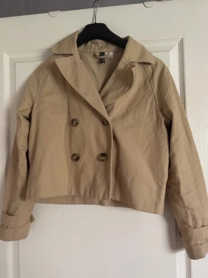 Kort beige trenchcoat från H&M - Stilren kort trenchcoat från H&M i beige bomullsmix. Jackan har dubbelknäppning framtill, bred krage och dekorativa knappar på ärmsluten. Klassisk design med en modern twist tack vare den korta modellen. Perfekt för dig som gillar minimalistisk och trendig stil.
