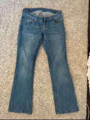 Blå bootcut jeans med klassisk look - Säljer ett par blå bootcut jeans med klassisk femficksdesign och normalhög midja. Jeansen har en snygg tvätt och subtila slitningar, samt raka ben som blir lite vidare nedtill. Perfekta för en avslappnad och trendig stil.