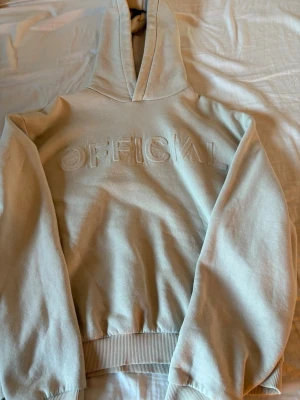 Beige hoodie från boohooMAN - Snygg beige hoodie från boohooMAN med broderad text 'OFFICIAL' på bröstet. Hoodien har huva, ribbade muddar och är gjord i mjukt bomullsmaterial med fleece på insidan. Perfekt för en avslappnad och trendig stil.