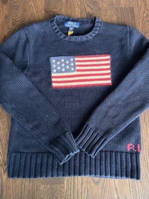 Mörkblå stickad tröja Polo Ralph Lauren - Mörkblå stickad tröja från Polo Ralph Lauren med amerikansk flagga broderad på bröstet och RL-broderi vid nederkanten. Tröjan har rund halsringning, långa ärmar och ribbstickade muddar. Perfekt för dig som gillar klassisk preppy stil.