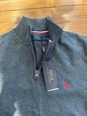 Grå halvzip tröja från Polo Ralph Lauren - HELT NY (!!!), Mörkgrå stickad tröja från Polo Ralph Lauren. Hör av er vid funderingar. Priset kan diskuteras något vid snabb affär!