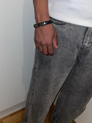 Svart flätat läderarmband med detaljer - Snyggt svart armband i flätat läder med flera silvriga metallringar och ett stilrent magnetlås. Perfekt accessoar för en modern och stilren look. Passar både till vardagsoutfits och mer dressade tillfällen.