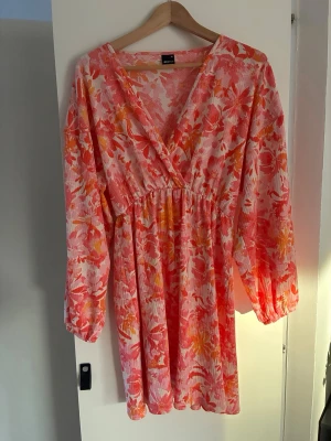 Blommig sommarklänning xs,  i rosa och orange - Superfin kort klänning med blommigt mönster i rosa, orange och vitt, från Gina. Klänningen har v-ringning, långa ärmar med resår vid handlederna och markerad midja. Perfekt för sommarens alla tillfällen! Storlek xs