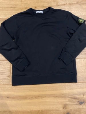 Svart sweater från Stone Island - Svart stickad tröja från Stone Island med klassisk rund halsringning och ribbade muddar. Tröjan har den ikoniska Stone Island-loggan på vänster ärm. Perfekt för en stilren och avslappnad look.