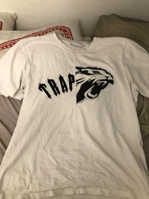 Vit t-shirt med tiger och TRAP-tryck - Cool vit t-shirt med svart broderad tiger och texten TRAP på bröstet. T-shirten har korta ärmar och en unik design där motivet är delat på mitten. Perfekt för dig som gillar streetwear och statement-plagg. Den är lite trasig där på tiger märket som ni ser annars är det inget fel på den.