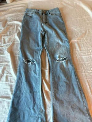 Ljusblå bootcut jeans med slitningar - Säljer ett par ljusblå bootcut jeans med slitna detaljer över knäna. Jeansen har klassisk femficksmodell, hög midja och vida ben. Perfekta för en avslappnad och trendig look. Materialet är mjuk denim i bomull.