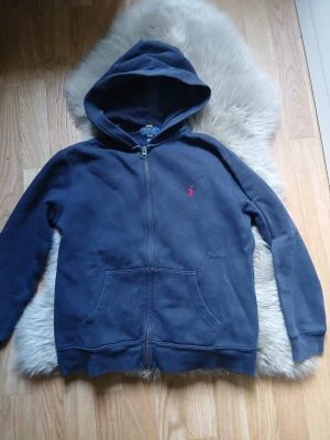 Marinblå hoodie från Polo Ralph Lauren - Marinblå hoodie från Polo Ralph Lauren med dragkedja och huva. Klassisk röd logga broderad på bröstet och två stora fickor framtill. Tillverkad i mjuk bomull, perfekt för en avslappnad stil. Skick 7/10