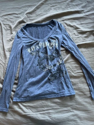 Blå långärmad topp med tryck - Snygg blå långärmad topp med v-ringning och coolt tryck framtill med text och detaljer som rosor och hjärtan. Perfekt för dig som gillar en edgy stil och vill sticka ut lite extra. Mjuk och skön att ha på sig.