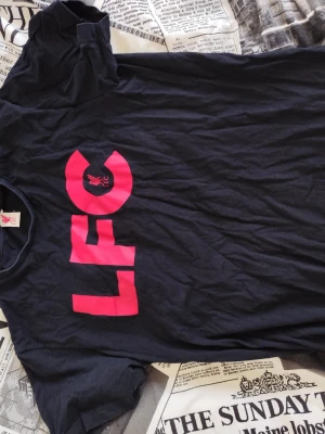 Svart LFC t-shirt med röd logga - Cool svart t-shirt från Liverpool FC med stor röd LFC-logga på bröstet. Klassisk passform och rund halsringning, perfekt för dig som vill visa ditt stöd för laget. Tillverkad i mjuk bomull som är skön att ha på sig hela dagen.