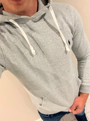 Grå hoodie från Ralph Lauren - Snygg ljusgrå hoodie från Ralph Lauren med klassisk broderad logga på bröstet. Hoodien har vita dragsnören, känguruficka och ribbade muddar. Perfekt för en avslappnad och stilren look.