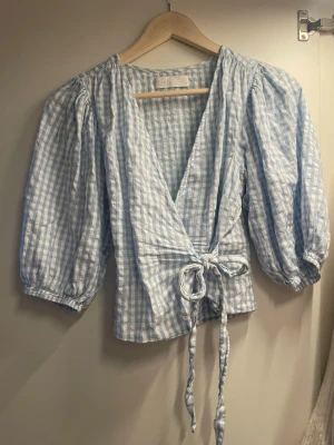 Blåvitrutig omlottblus med puffärm - Supersöt omlottblus i blåvitt rutigt tyg med puffiga trekvartsärmar och knytband i midjan. Djup v-ringning framtill och lätt strukturerat material som känns luftigt och fräscht. Perfekt för en trendig och avslappnad look.