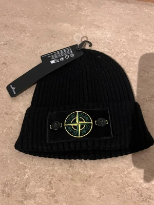 Svart ribbad mössa från Stone Island - Svart ribbstickad mössa från Stone Island med klassisk logotyp framtill på en patch med två knappar. Snygg och enkel design som passar till det mesta. Perfekt accessoar för kalla dagar.