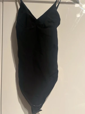 Svart body med v-ringning - Snygg svart body med smala axelband och v-ringad hals. Modellen är figurnära och har en lätt rynkning vid bysten. Materialet ser ut att vara stretchigt och bekvämt, perfekt för en stilren look.