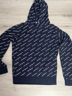 Marinblå hoodie från GANT med logga - Snygg marinblå hoodie från GANT med vit logotyp tryckt över hela tröjan. Klassisk huva med dragsko och ribbade muddar vid ärmslut och nederkant. Perfekt för en avslappnad och trendig stil.