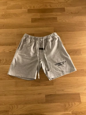 Grå shorts från Fear of God Essentials - Snygga grå shorts från Fear of God Essentials med svart logga på vänster ben. De har resår i midjan, dragsko och sidofickor. Perfekta för en avslappnad och trendig stil. Tillverkade i mjukt bomullsmaterial.