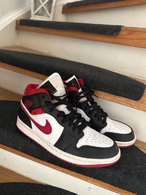 Air Jordan 1 Mid svart/röd/vit - Snygga Air Jordan 1 Mid sneakers i svart, vitt och rött läder. Klassisk siluett med svart swoosh, röda detaljer och perforerad tå. Skorna har svart snörning och ikoniska Jordan-loggor på sidan och plösen. Perfekta för dig som gillar streetwear och basketstil.