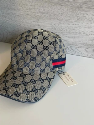 Gucci keps med GG-mönster och rand - Snygg keps från Gucci i beige och marinblått GG-mönster. Kepsen har en klassisk böjd skärm, justerbar rem bak i läder och en ikonisk röd-blå rand på sidan. Tillverkad i canvasmaterial med läderdetaljer. Perfekt för dig som gillar lyxiga accessoarer.