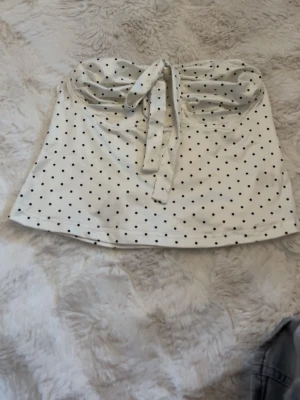 Vit prickig tubtopp med knyt - Snygg vit tubtopp med svarta prickar och knyt framtill. Toppen är axelbandslös och har en bandeau-form som ger en trendig look. Perfekt att matcha med jeans eller kjol för en cool outfit. Den står att de är xs men skulle säga att den passa s