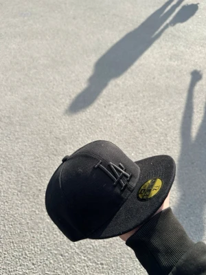 Svart LA-keps från New Era - Snygg svart keps från New Era med LA-logga framtill i svart brodyr. Klassisk flat peak och guldigt klistermärke på skärmen. Perfekt för dig som gillar streetwear och vill ha en stilren look.