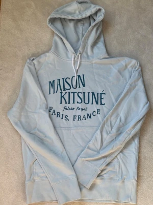 Maison Kitsuné ljusblå hoodie - Snygg ljusblå hoodie från Maison Kitsuné med tryck i mörkblått framtill där det står 'Maison Kitsuné Palais Royal Paris, France'. Hoodien har huva med vita dragsnören och en stor magficka. Perfekt för en chill och trendig look.