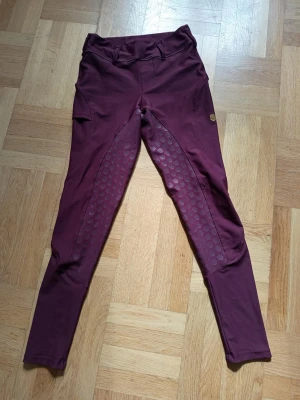 Vinröda ridbyxor med silikonprint - Snygga vinröda ridbyxor från Maya delorez med helskodd silikonprint för extra grepp. Byxorna har hög midja, smal passform och praktisk ficka på låret. Perfekta för dig som gillar stilrena och funktionella ridkläder.