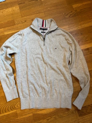 Grå halvzip-tröja Tommy Hilfiger - Snygg grå halvzip-tröja från Tommy Hilfiger med liten broderad logga på bröstet. Tröjan har ribbade muddar, hög krage med dragkedja och insida med röd, vit och marinblå rand. Tillverkad i mjuk bomull. Använd 2 gånger!