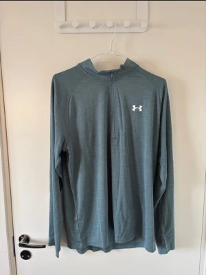 Grön långärmad träningströja Underarmour - Oanvänd, ny med prislapp. Nypris:549kr Snygg grön långärmad träningströja från Under Armour med halv dragkedja och vit logga på bröstet. Tillverkad i ett lätt och tekniskt material som andas, perfekt för löpning eller gym. Regular fit och bekväm passform.