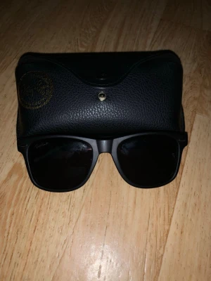 Ray-Ban Wayfarer svarta solglasögon - Snygga svarta Ray-Ban Wayfarer solglasögon med klassisk form och mörka glas. Kommer med original svart fodral i skinnimitation med guldig Ray-Ban-logga. Perfekt accessoar för en stilren look.