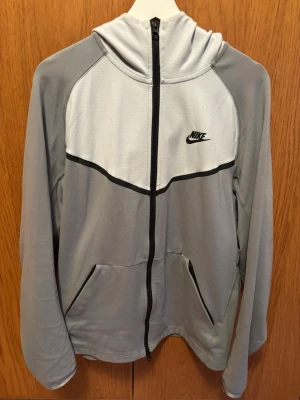 Grå nike tech kofta - Grå Nike tech kofta med svart logga på bröstet och svarta detaljer längs dragkedjan och bröstet. Tröjan har huva, två fickor framtill och är tillverkad i ett mjukt material som känns skönt mot huden. Perfekt för en sportig och avslappnad stil.