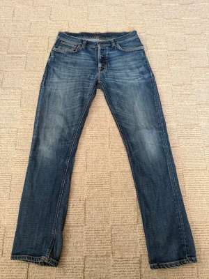 Blå jeans från Nudie Jeans - Säljer ett par klassiska blå jeans från Nudie Jeans med snyggt slitna detaljer och raka ben. Jeansen har fem fickor, orangea sömmar och är gjorda i mjuk denim. Perfekta för en avslappnad och trendig stil. De har ett hål på baksidan vid fickan men går att laga gratis på en Nudie butik. Storlek W30 L32 mått: längd 103 cm midja platt 40 cm och benöppningen 18 cm