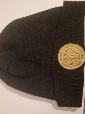 Svart mössa från Mitchell & Ness - Svart ribbstickad mössa med uppvikt kant från Mitchell & Ness. Broderad logotyp i guld på sidan och ett runt tygmärke framtill. Perfekt för kalla dagar och snygg streetstil.