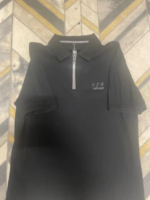 Svart polotopp med dragkedja - Svart polotopp med kort ärm och silverfärgad dragkedja framtill. Toppen har en klassisk krage och en diskret logga med texten 'OZ' på bröstet. Perfekt för en stilren och sportig look.