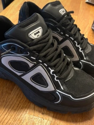 Svarta sneakers med vita detaljer - Snygga svarta sneakers med vita och grå detaljer samt svart mesh och läder. Skorna har en chunky sula och coola kontrastmönster på sidorna. Perfekta för dig som vill sticka ut med ett exklusivt streetwear-utseende.