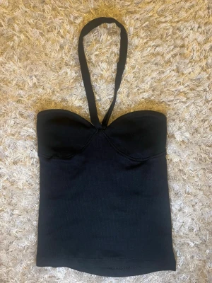 Svart halterneck tubtopp - Säljer en svart tubtopp med halterneckband och lätt vadderade kupor. Toppen har en tight passform och är tillverkad i ett stretchigt ribbat material. Perfekt för dig som vill ha en enkel men snygg topp till sommaren.
