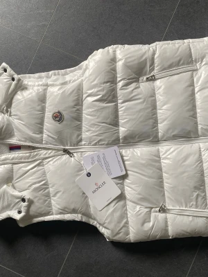 Vit dunväst från Moncler - Snygg vit dunväst från Moncler med klassisk quiltad design och dragkedja framtill. Västen har två fickor med dragkedja och Moncler-logga på bröstet. Tillverkad i glansigt material och fylld med dun för extra värme. Perfekt för lager-på-lager-stil.