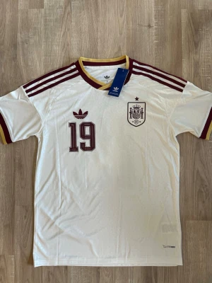 Spanien Lamine Yamal VM-2026 Fotbollströja - Vit Spanien fotbollströja med vinröda detaljer, nummer 19 och Lamine Yamal på ryggen. Tröjan har klassiska Adidas-ränder på axlarna, klubbmärke och CLIMACOOL-material för bra ventilation. Perfekt för dig som älskar fotboll och vill stötta Spanien. HELT NY