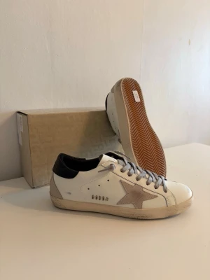 Golden Goose vita sneakers med stjärna - Golden Goose sneakers i vitt läder med grå mockastjärna på sidan, grå skosnören och svart hälparti. Sulan är beige och har en vintage-look. Ikonisk design med låg profil och detaljer i metall. Perfekta för en trendig streetstyle.