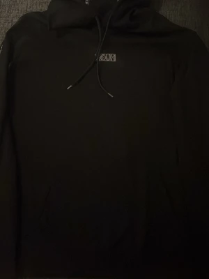 Svart hoodie från Hugo Boss - Snygg svart hoodie från Hugo Boss med liten logga på bröstet och dragsko i huvan. Klassisk modell med känguruficka framtill och ribbade muddar. Perfekt för en stilren och avslappnad look.