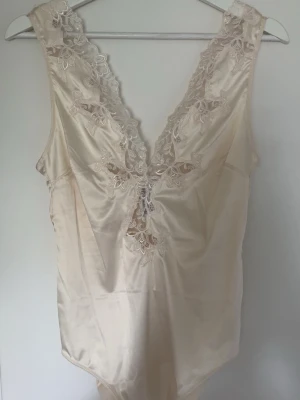 Krämfärgad spetsbody med djup rygg - Elegant body i krämfärgad satin med v-ringad rygg och vackra spetsdetaljer längs kanterna.