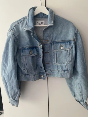 Ljusblå cropped jeansjacka från NA-KD - Trendig ljusblå jeansjacka från NA-KD i croppad modell. Jackan har klassisk krage, två bröstfickor med lock och silverfärgade knappar. Tillverkad i jeansmaterial. Säljer då jag inte fått användning för den☺️