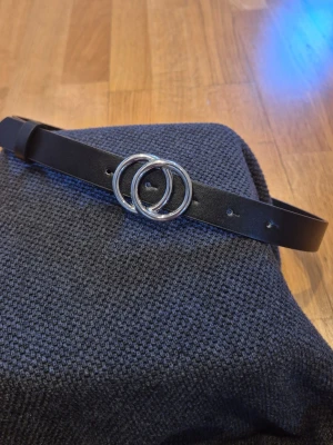 Svart bälte med silverfärgat spänne från Lindex - Snyggt svart bälte med ett stilrent, dubbelt silverfärgat ring-spänne från Lindex. Bältet är smalt och passar perfekt till jeans eller kjol. Materialet känns mjukt och flexibelt, och designen är enkel men trendig.