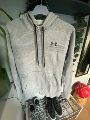  Under Armour - Snygg grå hoodie från Under Armour med svart logga på bröstet. Hoodien har huva med svarta dragsnören och en stor magficka framtill. Tillverkad i mjukt och bekvämt material, perfekt för chill eller träning.