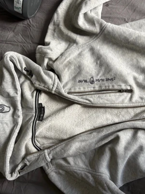 Grå hoodie från Sail Racing - Snygg grå hoodie från Sail Racing med dragkedja framtill och broderad logga på bröstet. Hoodien har lite tecken på användning som visas på bilderna, annars inga problem 