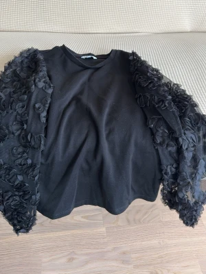 Svart topp med volangärmar - Svart topp med unika, transparenta volangärmar dekorerade med blommiga detaljer. Rund halsringning och rak passform. Perfekt för dig som vill sticka ut med en cool och trendig look.