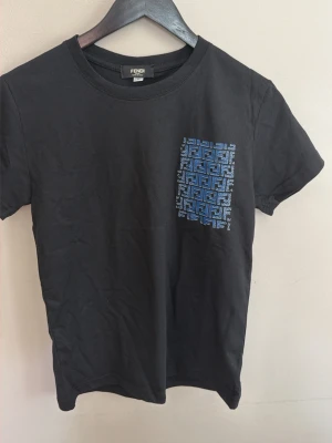 Svart Fendi t-shirt med blått tryck - Snygg svart t-shirt från Fendi med ett blått grafiskt tryck på bröstet. Klassisk rund halsringning och korta ärmar. Tillverkad i mjuk bomull som känns skön mot huden. Perfekt för dig som gillar stilrena plagg med en twist.