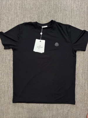 Svart t-shirt från M - Snygg svart t-shirt från M med klassisk rund hals och diskret M-logga på bröstet. T-shirten har en enkel och stilren design, perfekt för en clean look. Tillverkad i mjuk bomull för skön komfort.